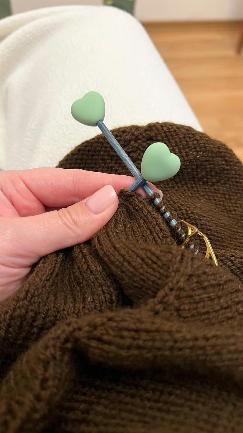 Una mano lavora a maglia con filato marrone e un ferro a forma di cuore.